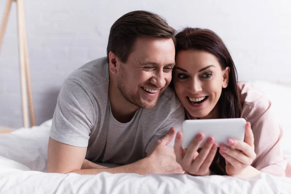 Mujer sonriente sosteniendo el teléfono celular en primer plano borroso cerca del marido en el dormitorio - foto de stock