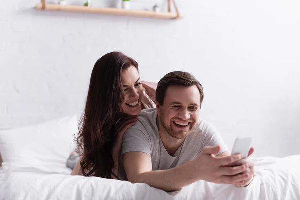 Uomo sorridente utilizzando smartphone in primo piano sfocato vicino a moglie allegra sul letto — Foto stock