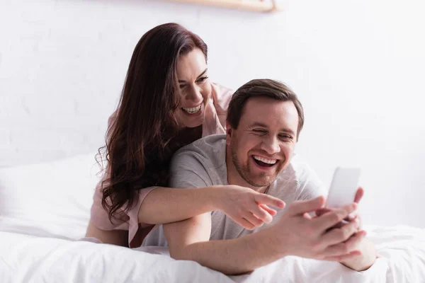 Mulher alegre apontando com o dedo para o smartphone em primeiro plano borrado perto do marido no quarto — Fotografia de Stock