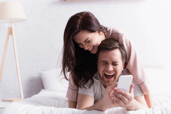 Rindo homem segurando celular em primeiro plano borrado perto da esposa na cama de manhã — Fotografia de Stock