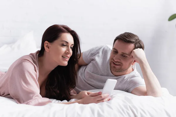 Brunette woman holding smartphone near husband in bedroom — Stock Photo