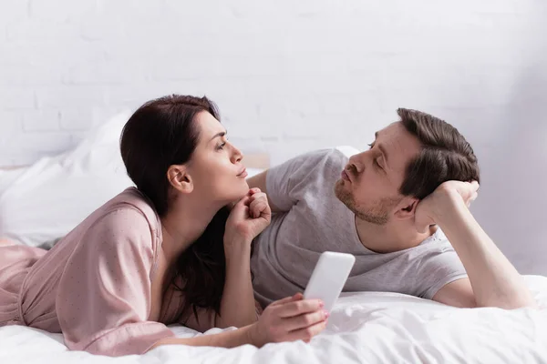 Femme avec smartphone boudant lèvres près du mari sur le lit — Photo de stock