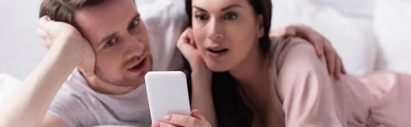 Smartphone in hand of woman lying near man on bed on blurred background, banner — Stock Photo