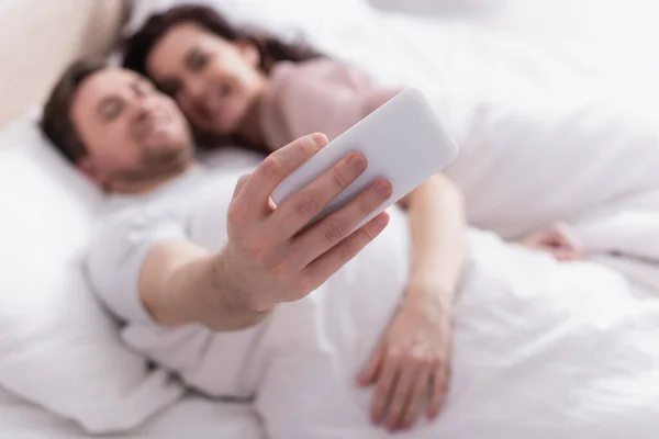 Smartphone in hand of man taking selfie near wife in bed on blurred background — Stock Photo
