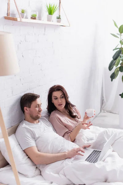 Woman with cup talking to husband using laptop in bed — Stock Photo