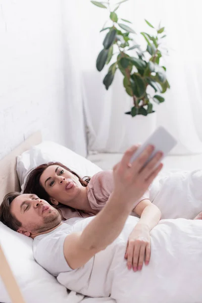 Adult couple sticking out tongues while taking selfie in bed — Stock Photo