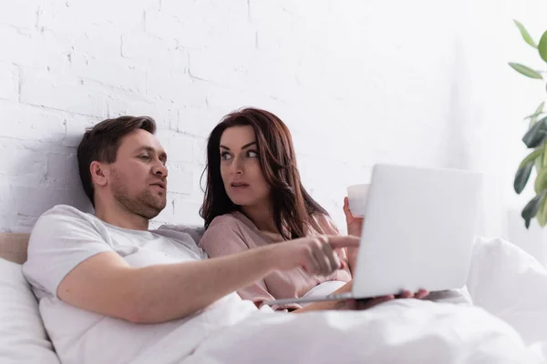 Man pointing with finger at laptop near wife with cup on bed — Stock Photo