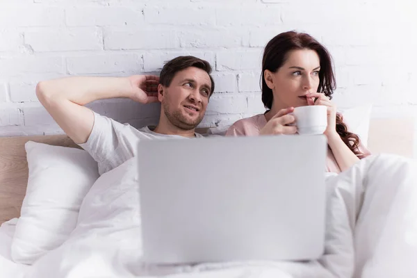 Femme stressée tenant une tasse de café près du mari et de l'ordinateur portable au premier plan flou sur le lit — Photo de stock