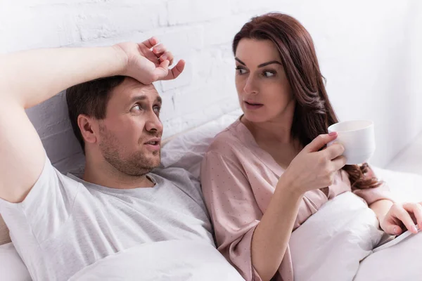 Mujer con taza de café y teléfono inteligente mirando al marido en la cama - foto de stock