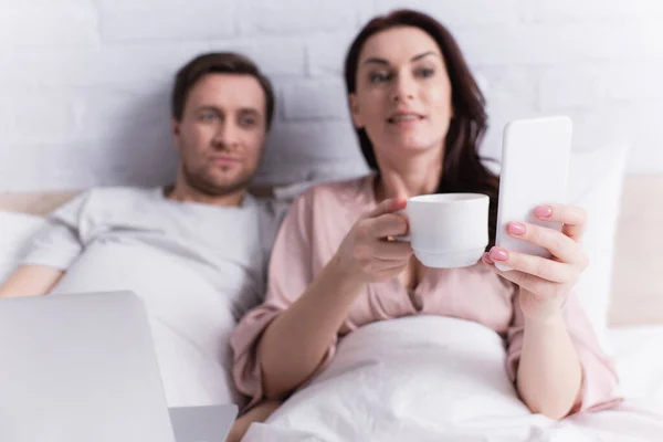 Smartphone in hand of woman with coffee lying near laptop and husband on bed — Stock Photo