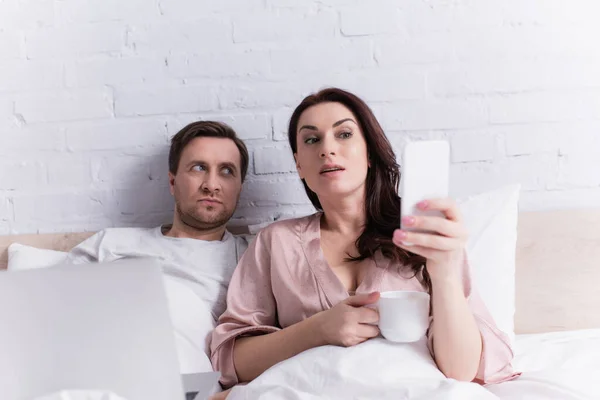 Woman using cellphone and holding cup near husband with laptop on bed — Stock Photo