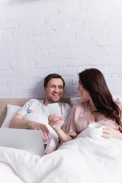 Smiling woman with cup showing smartphone to husband with laptop on bed — Stock Photo