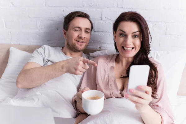 Cheerful woman with coffee holding smartphone near husband pointing with finger and laptop in bedroom — Stock Photo