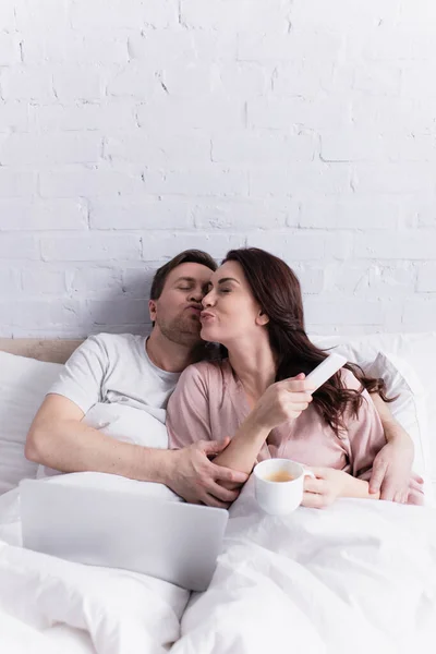 Женщина с кофе и смартфоном целует мужа рядом с ноутбуком на кровати — стоковое фото