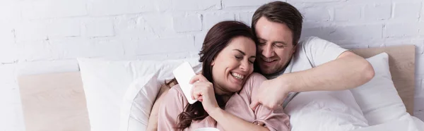 Усміхнений чоловік лоскоче дружину зі смартфоном на ліжку, банер — стокове фото