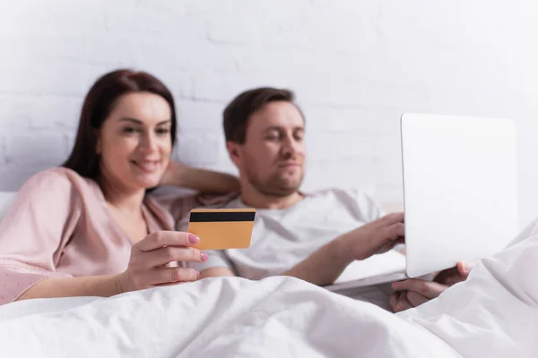 Cartão de crédito na mão de mulher deitada perto do marido com laptop no fundo embaçado na cama — Fotografia de Stock