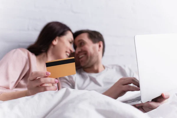 Tarjeta de crédito en la mano de la mujer adulta sonriendo al marido con el ordenador portátil sobre fondo borroso en el dormitorio - foto de stock