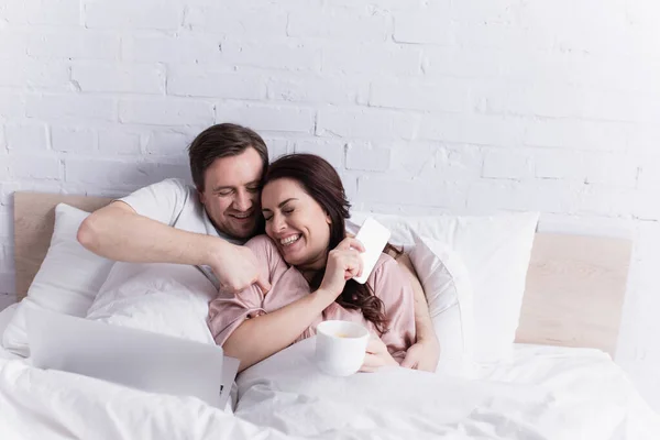 Homem cócegas esposa alegre com smartphone e café perto de laptop na cama — Fotografia de Stock