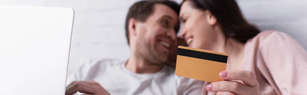 Credit card in hand of woman smiling near husband with laptop on blurred background, banner — Stock Photo