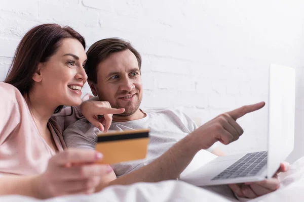 Sonriente hombre apuntando a la computadora portátil cerca de la esposa con tarjeta de crédito en primer plano borroso en la cama - foto de stock