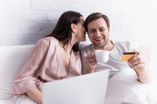 Woman whispering to smiling husband with credit card and coffee near laptop on blurred foreground on bed — Stock Photo