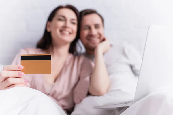 Credit card in hand of blurred woman near husband and laptop on bed — Stock Photo