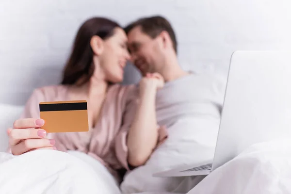 Laptop near woman holding credit card and looking at husband on blurred background on bed — Stock Photo
