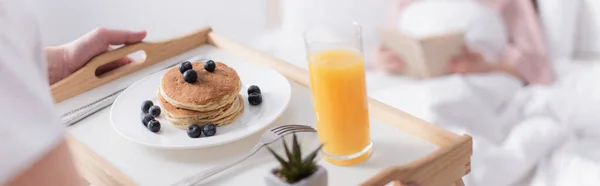 Vista recortada del hombre sosteniendo panqueques y vaso de jugo de naranja en bandeja cerca de la esposa borrosa en el dormitorio, pancarta - foto de stock