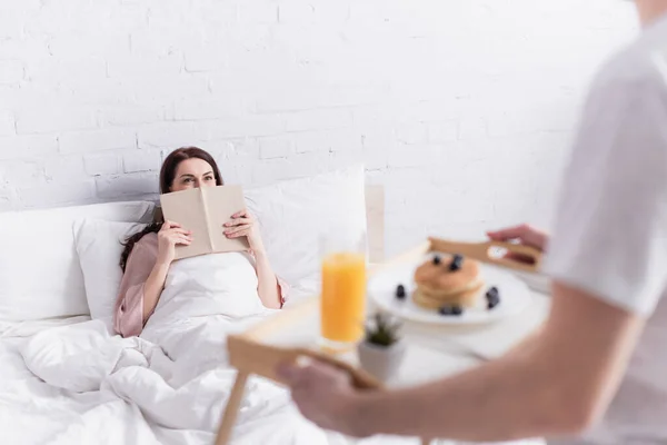 Frau hält Buch neben Ehemann mit Frühstück auf verschwommenem Vordergrund im Schlafzimmer — Stockfoto