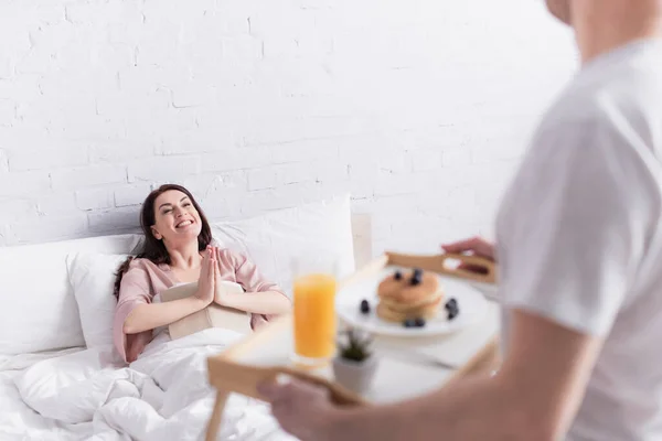 Mujer sonriente con libro mostrando gesto por favor cerca de marido borroso con desayuno en el dormitorio - foto de stock