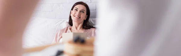 Mujer sonriente con libro mirando al marido borroso con panqueques en el dormitorio, pancarta - foto de stock