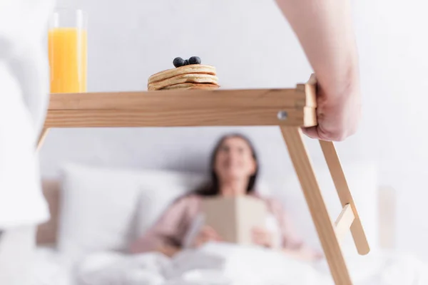 Homem segurando panquecas e suco de laranja na bandeja de madeira perto da esposa na cama no fundo borrado — Fotografia de Stock