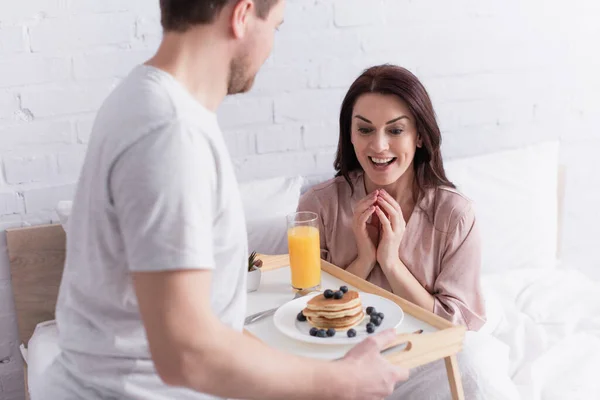 Mulher feliz olhando para panquecas saborosas e suco de laranja na bandeja perto do marido no quarto — Fotografia de Stock