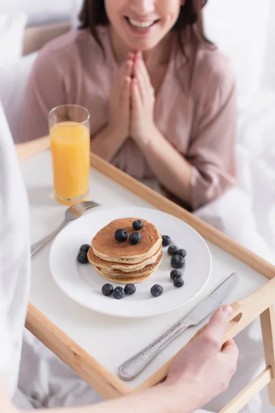 Vista cortada do homem segurando bandeja de café da manhã com panquecas e bagas perto de esposa sorridente no fundo borrado na cama — Fotografia de Stock