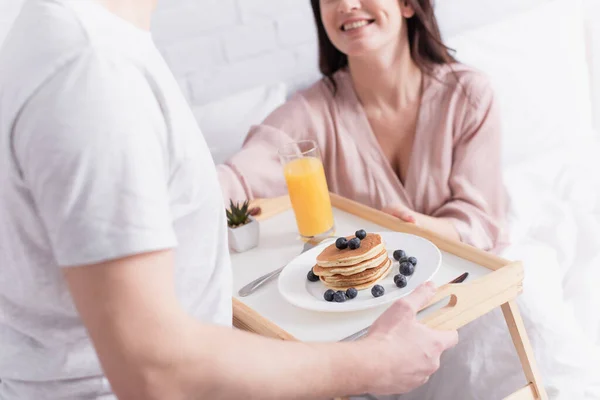 Cropped view of delicious pancakes and orange juice on tray near couple on blurred background in bedroom — Stock Photo