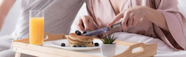Cropped view of woman cutting pancakes near orange juice and husband on bed, banner — Stock Photo