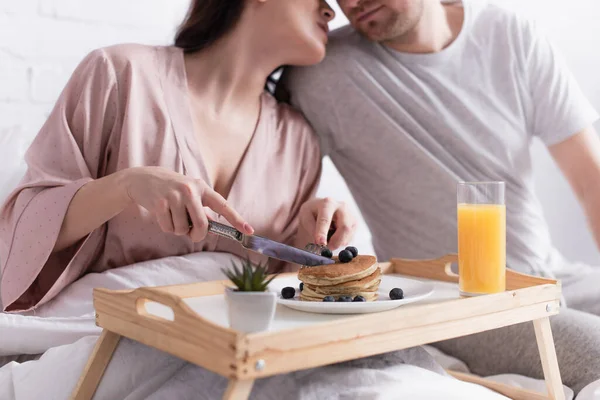 Cropped view of woman holding cutlery near pancakes and husband on blurred background on bed — Stock Photo