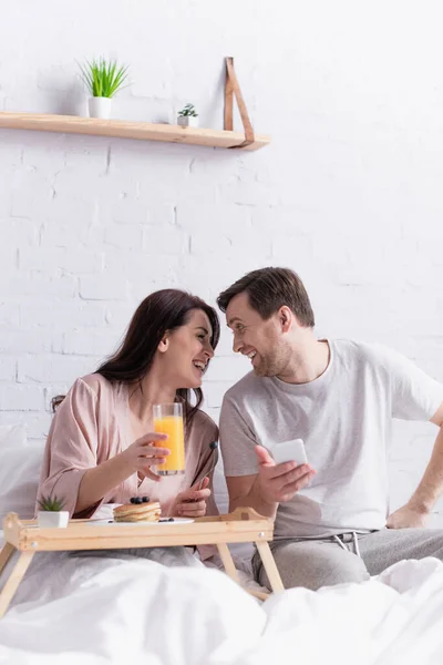 Femme souriante tenant du jus d'orange près du petit déjeuner et mari avec smartphone — Photo de stock