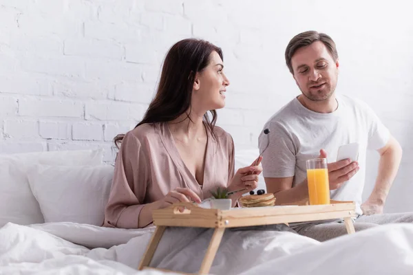 Mulher olhando para o marido com smartphone perto do café da manhã em primeiro plano desfocado na cama — Fotografia de Stock