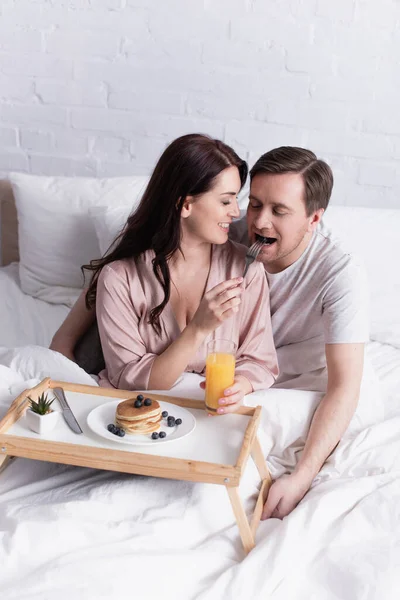 Mujer sonriente alimentando marido cerca de panqueques y jugo de naranja en la cama - foto de stock