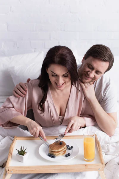 Sourire homme étreignant femme coupe crêpe près de jus d'orange sur plateau sur le lit — Photo de stock