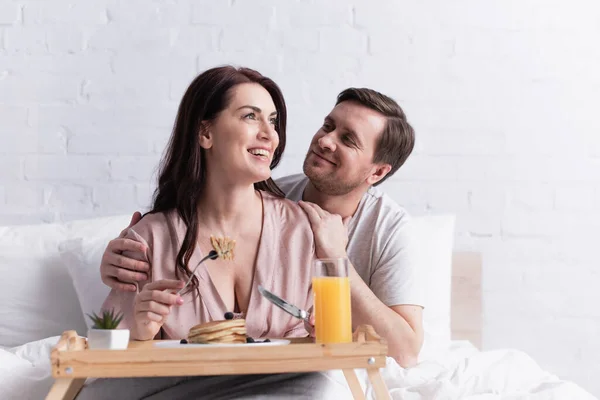 Мужчина обнимает жену со столовыми приборами рядом с размытым завтраком на кровати — стоковое фото