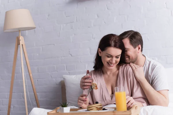 Uomo che abbraccia donna felice con forchetta vicino a colazione e succo d'arancia sul letto — Foto stock