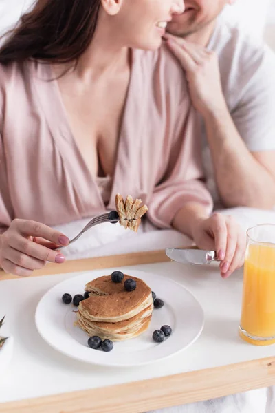 Cropped view of woman holding cutlery near pancakes with berries, orange juice and husband on blurred background — Stock Photo