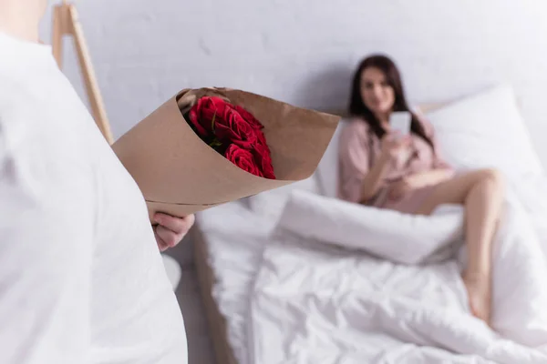 Man holding bouquet of roses near wife on bed on blurred background — Stock Photo