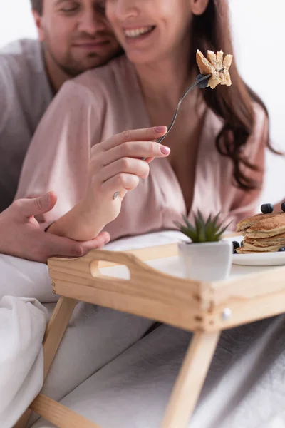 Vista ritagliata di frittelle sulla forchetta in mano della donna sorridente vicino al marito su sfondo sfocato sul letto — Foto stock