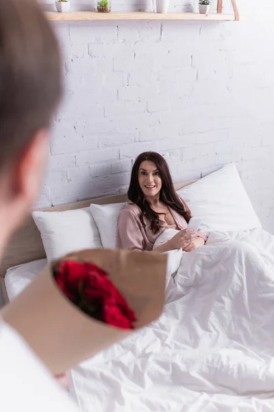 Усміхнена жінка зі смартфоном дивиться на чоловіка з трояндами на розмитому передньому плані в спальні — стокове фото