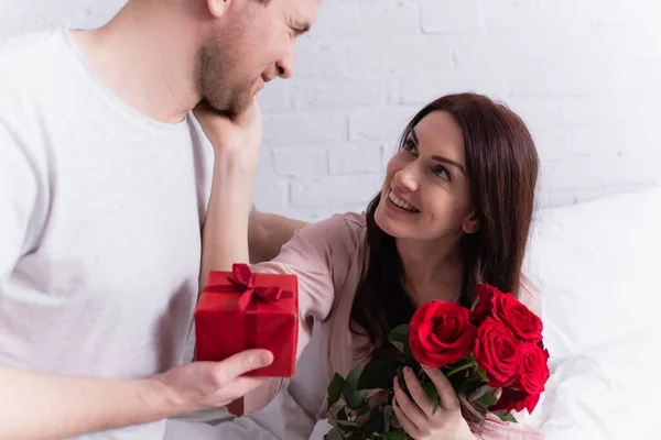 Smiling woman with roses touching face of husband with present on bed — Stock Photo