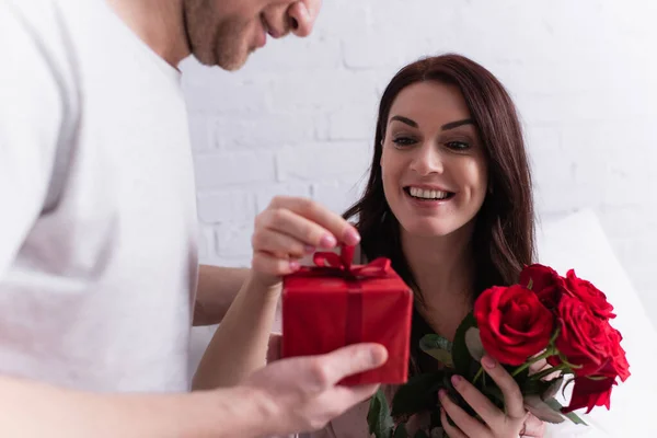 Cheerful woman with roses holding bow of present near husband on blurred foreground — Stock Photo