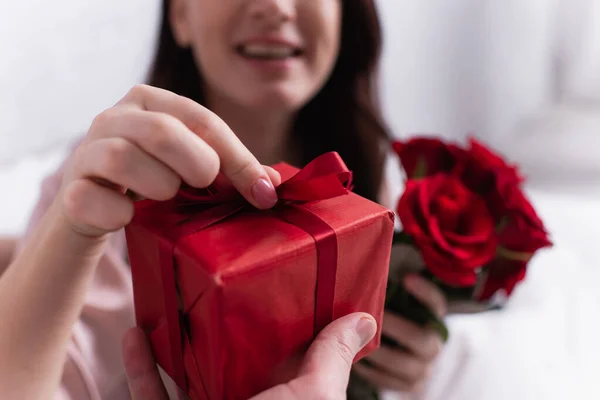 Vista cortada do homem segurando presente perto da esposa com flores no fundo borrado — Fotografia de Stock